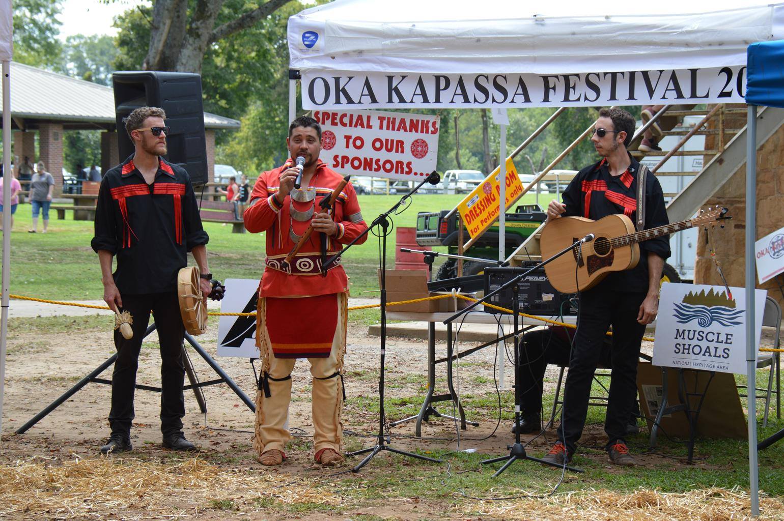 Three American Indian men standing under a tent playing native musical instruments.