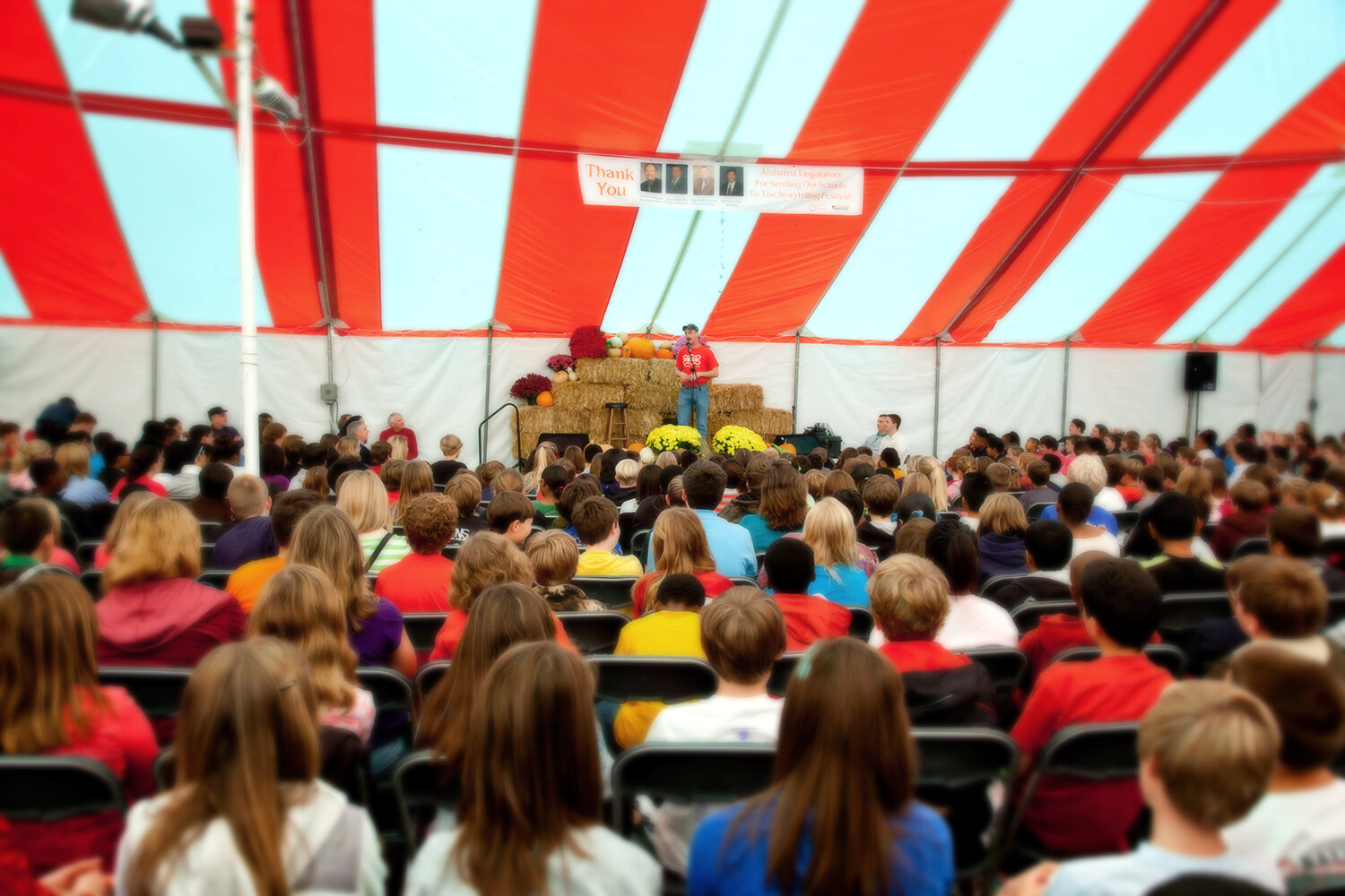 An audience seated under a large red-and-white striped tent listening to someone speak. Decorations include pumpkins and hay bales.