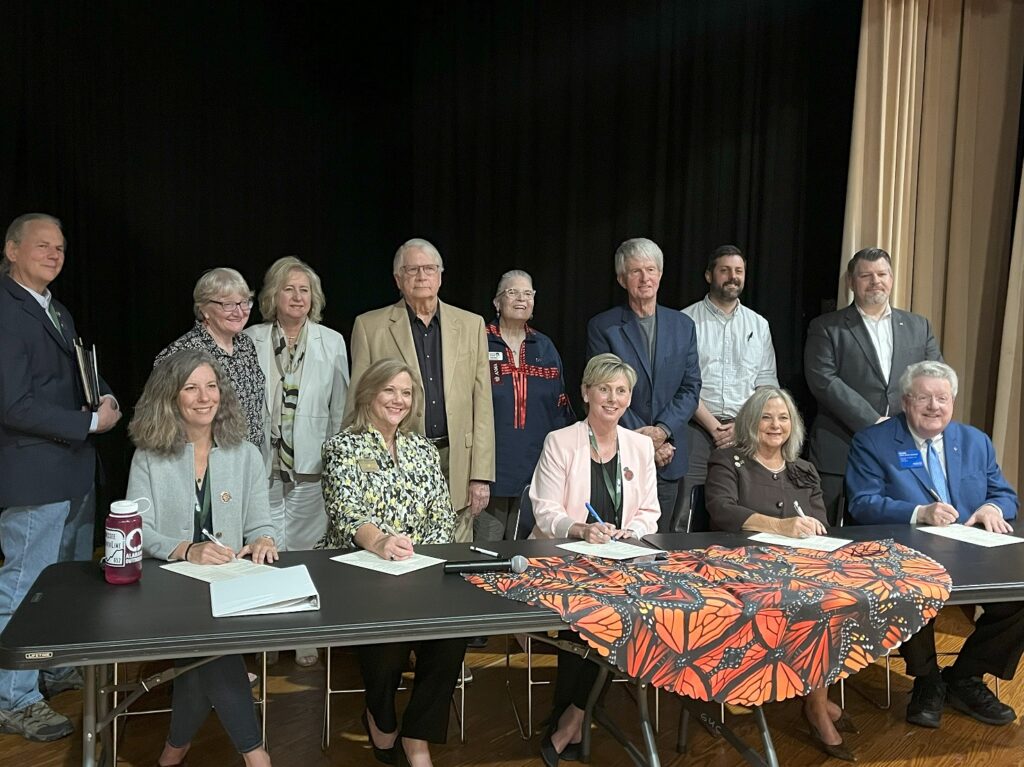 Four women and one man dressed in business attire sit on stage behind a black table draped with a monarch butterfly-print tablecloth. They are each signing a document on the table in front of them while smiling facing the camera. A group of eight similarly dressed individuals pose standing behind them in show of support.