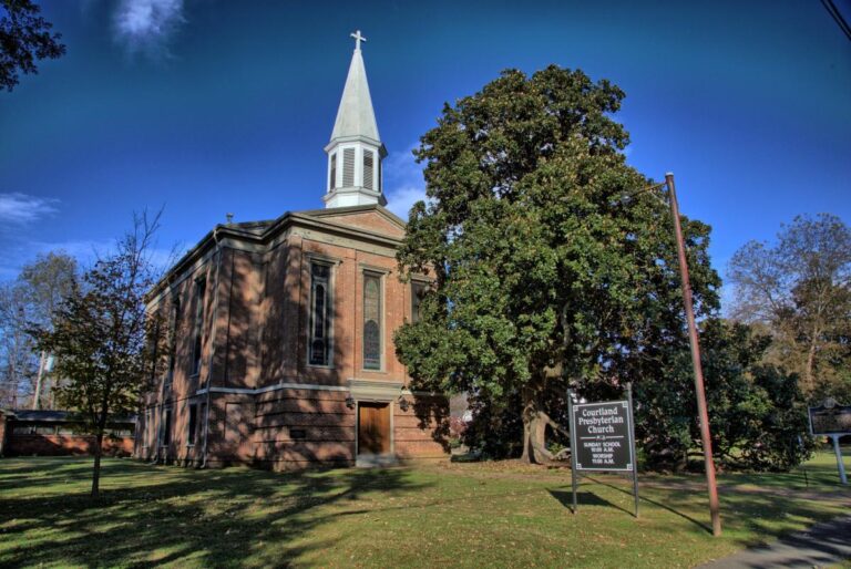 Photo of an old brick church with a white steeple on a summer day with blue skies and fully leafed-out green trees.