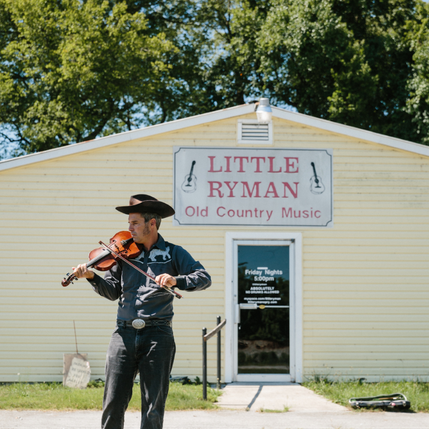 A man wearing country-music attire playing a fiddle in front of the Little Ryman music hall.